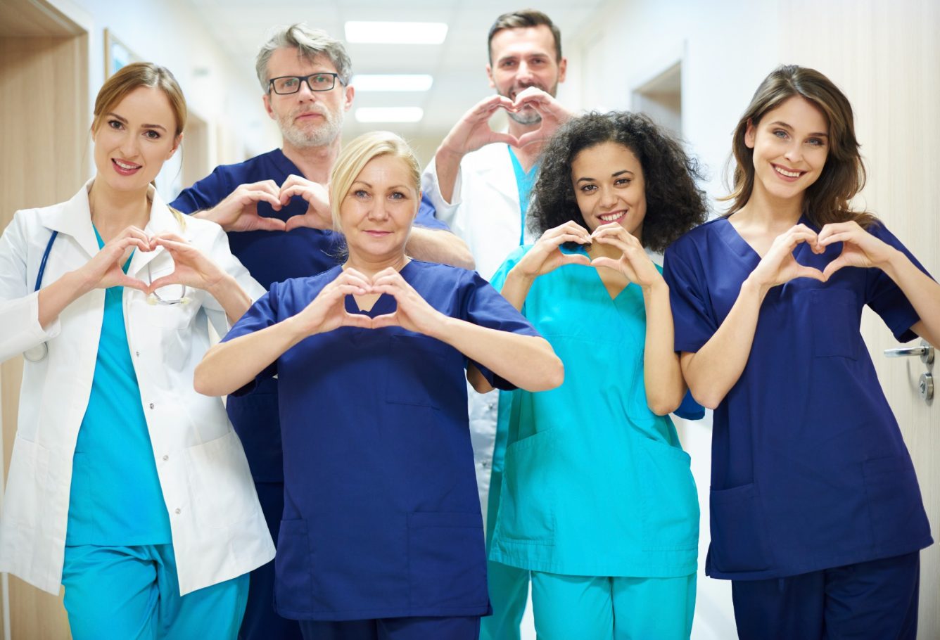 Healthcare team making heart shapes with their hands, smiling in a hospital hallway.